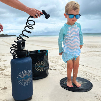 Photograph of a young child in a blue striped rashguard and mirrored sunglasses being rinsed by a dark blue Beach Soul portable shower on a black textured mat on a white sandy beach with distant boats