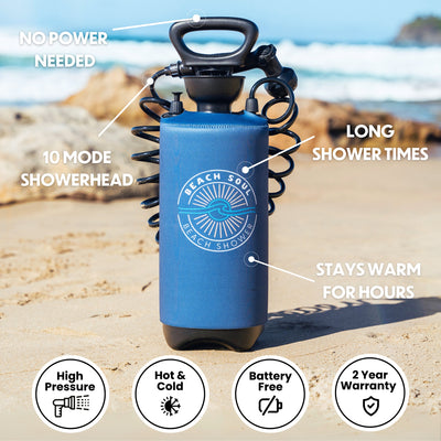 A blue Beach Soul portable shower with a black handle and coiled 10-mode showerhead stands on a sandy beach featuring ocean waves and rocky cliffs, prominently advertising No Power Needed and Stays Warm for Hours