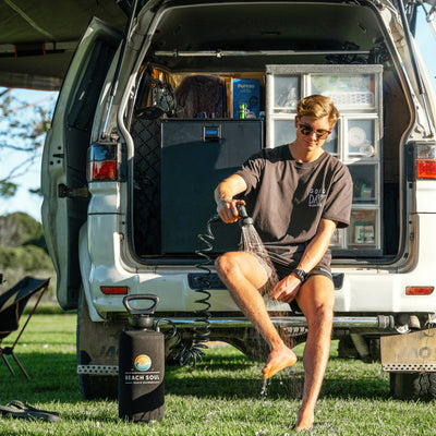 A young man in sunglasses and a 'GOOD DAY TO EXPLORE' t-shirt sits on a white SUV tailgate, showering his bare feet with a 'BEACH SOUL' portable shower on green grass, featuring an 'Adventure Kings' fridge and camping storage in the vehicle's rear