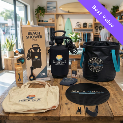 A collection of Beach Soul brand beach and outdoor products displayed on a rustic wooden table in a bright surf shop with surfboards prominent in the background, featuring a black 10L portable shower, a black collapsible bucket with vibrant teal handles, and a beige canvas tote bag, all adorned with the brand's colorful sun and wave logos, beneath a purple Best Value banner