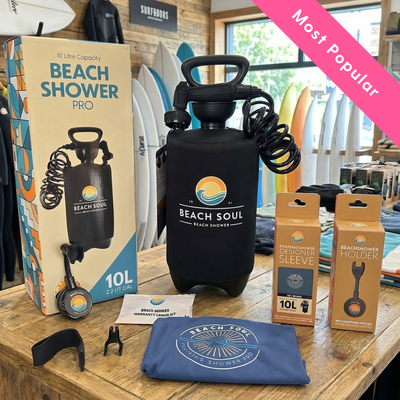 Beach Soul Beach Shower Pro 10L portable shower kit with accessories displayed on a rustic wooden table in a surf shop featuring several colorful surfboards and a Most Popular pink banner