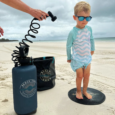 Photograph of a young boy in a blue wavy rash guard and reflective sunglasses getting rinsed by a portable Beach Soul shower on a wide sandy beach with tire tracks under an overcast sky
