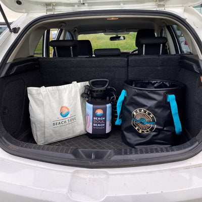 Beach Soul branded beach shower tote bag, portable pressure shower, and collapsible bucket with bright turquoise handles neatly packed in the trunk of a white car