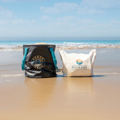 A black Beach Soul collapsible bucket with teal handles and a white fabric Beach Soul bag rest on wet sand at the ocean's edge featuring vibrant blue waves and a clear sky