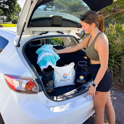 Photograph of a young woman in a green sports bra and black shorts packing a white car trunk with a bright turquoise fringed towel, alongside a white "Beach Soul" tote bag and black portable shower, set outdoors with lush green foliage