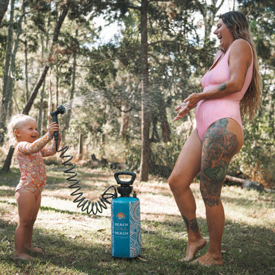 A joyful mother in a pink swimsuit with extensive leg tattoos and a laughing toddler in a floral rashguard spraying water from a blue Beach Soul portable shower in a sunny forest clearing