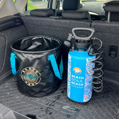 A black collapsible bucket with turquoise handles and a gold and turquoise "Beach Soul" logo sits beside a blue "Beach Soul Beach Shower" bottle featuring a white and orange mandala pattern and a coiled black hose, all resting on a rubber mat in a car trunk