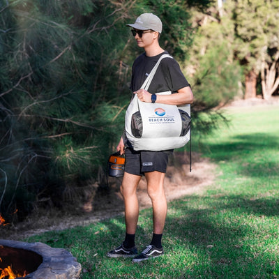 A young man wearing a grey cap, sunglasses, black shorts, and Vans sneakers stands on green grass next to a stone fire pit with visible flames, holding an orange and grey portable water container and a large white tote bag featuring the colorful Beach Soul logo