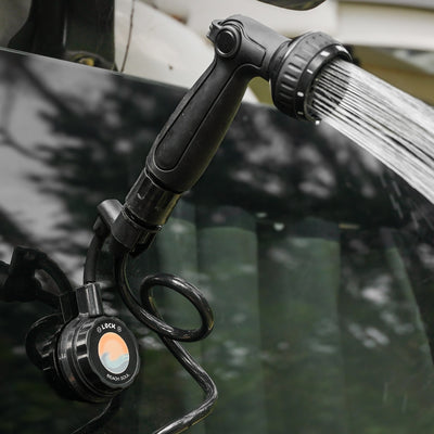 A black portable outdoor shower head attached to a dark vehicle with a coiled hose and a colorful Beach Soul logo sprays water from its multi-setting nozzle