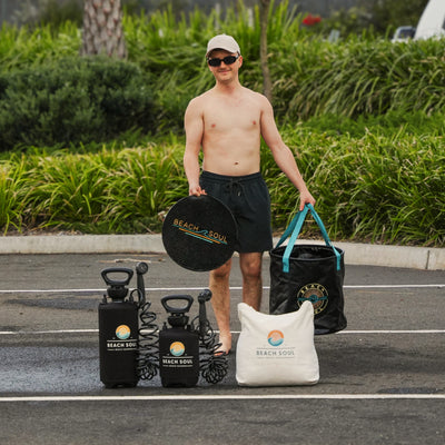 A shirtless man in a beige cap and sunglasses stands in a parking lot displaying Beach Soul branded portable beach showers a black collapsible bucket with teal handles a black textured drying mat and a white tote bag against a backdrop of vibrant green foliage