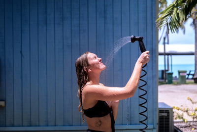 Photograph of a smiling young woman with wet blonde hair and a freckled face showering outdoors against a blue wooden wall, revealing a sun tattoo on her torso, with blurred palm trees and the ocean in the background