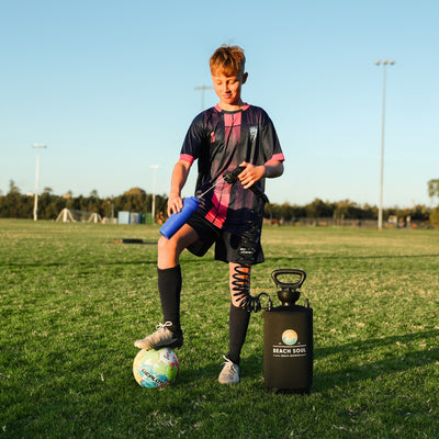 Photograph of a young male soccer player in a dark blue and pink jersey with his foot on a colorful soccer ball on a grassy sports field, holding a blue water bottle connected by a coiled hose to a black "Beach Soul" portable shower system under stadium lights