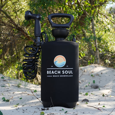 A black portable Beach Soul shower unit featuring a coiled hose and a colorful sunset and wave logo stands on sandy ground amidst green foliage