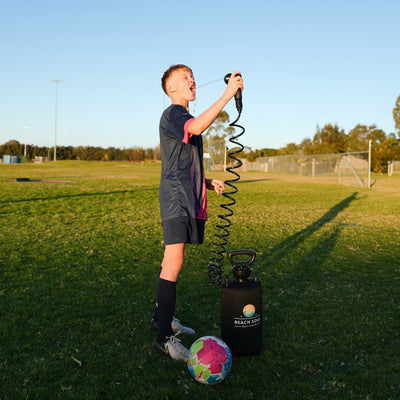 A young boy in a navy and pink soccer uniform takes a refreshing drink from a black Beach Soul portable shower on a bright green sports field, with a vibrant multi-colored soccer ball at his feet and a distant soccer goal visible