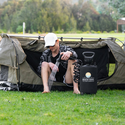 Photograph of a man in a white cap and sunglasses, wearing a plaid shirt, using a black Beach Soul portable shower to wash his bare feet while seated in the opening of a khaki swag tent on a green grassy campsite