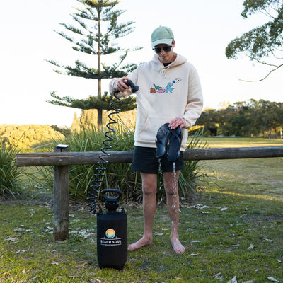 Photograph of a man outdoors cleaning black flip-flops with a portable Beach Soul Beach Shower, wearing a light hoodie with colorful dinosaur graphics, a green striped cap, and sunglasses, standing barefoot on grass next to a wooden fence and tall pine trees