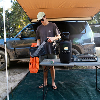A man in a Corona Extra t-shirt and green cap rinses a black pan using a Beach Soul portable shower system at an outdoor campsite beside a dirty grey SUV with orange recovery tracks and a camping stove on a grey table