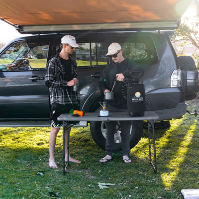 Photograph of two men at a sunny outdoor camp setup, one in a plaid shirt barefoot holding a drink, the other filling a pot with a Beach Soul portable shower system on a small blue camping stove next to a dark SUV with an extended awning
