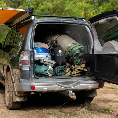 Photograph of a dark grey SUV with an orange side awning deployed and its open trunk overflowing with green camping gear including a black 'Beach Soul' portable shower, on a muddy dirt track surrounded by trees