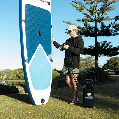 A young man in a beige cap and black hoodie cleans a blue and white geometric stand-up paddleboard with a black BEACH SOUL portable shower in a sunny grassy park by a calm lake with a tall pine tree
