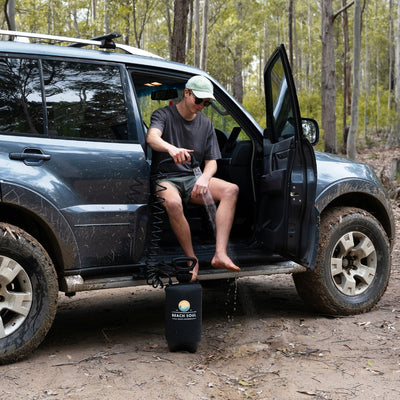A man sitting in the open door of a muddy SUV in a forest rinses his bare leg with a black Beach Soul portable shower surrounded by tall trees