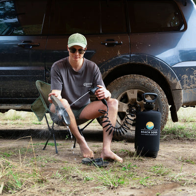 Photograph of a man wearing a green corduroy cap and Corona Extra t-shirt rinsing his bare foot with a black Beach Soul portable shower, sitting beside a muddy dark SUV in an outdoor setting