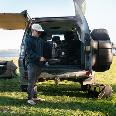 Photograph of a man in a white cap and dark hoodie washing a pan with water from a portable "BEACH SOUL" shower system at the back of a grey SUV parked on green grass by a calm body of water, a yellow and black cooler bag nearby