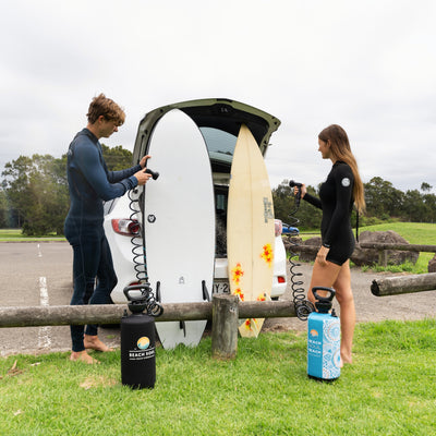 Two surfers in wetsuits, a man and a woman, rinse their surfboards, one white with a geometric pattern and one yellow with orange flowers, using a black and a blue mandala-patterned Beach Soul portable shower beside an open car boot in a grassy park