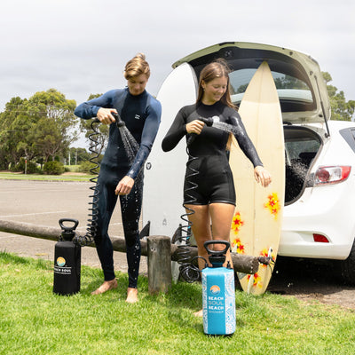 Photograph of two surfers in wetsuits rinsing off with black and turquoise "Beach Soul Beach Shower" portable units in a grassy beach parking lot, next to a white car and two surfboards, one decorated with red and yellow stars