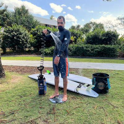 A bearded man in a black wetsuit and navy striped board shorts uses a portable "Beach on the Beach" sprayer to rinse off, standing on a circular patterned mat with two white surfboards and a black folding bucket on the grassy ground