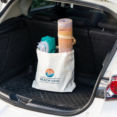 Photograph of a white Beach Soul tote bag featuring an orange sun and blue wave logo in a car trunk, holding a rolled sunset-striped mat and a teal fringed towel on a black textured mat