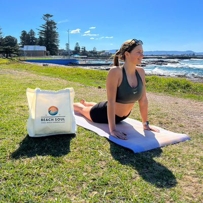 Photograph of a young woman in a green Nike sports bra performing upward-facing dog yoga on a purple mat in a grassy coastal park overlooking a blue ocean pool and rocky shoreline with crashing waves, next to a white Beach Soul tote bag under a clear blue sky