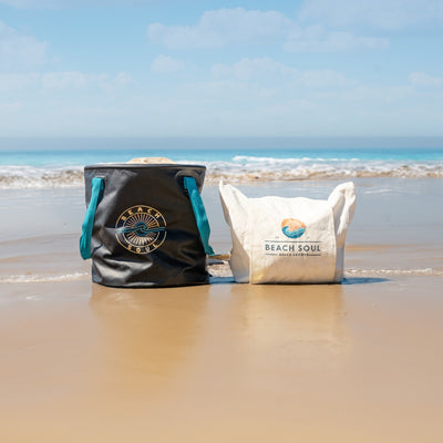 A black Beach Soul bucket bag with teal straps and a gold sun logo sits next to a white Beach Soul canvas tote with a blue wave logo on a wet sandy beach under a blue sky with ocean waves