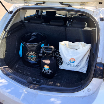 Beach Soul branded beach gear in the open trunk of a white car including a black collapsible bucket with blue handles, a black portable shower with a coiled hose, and a white canvas tote bag, all featuring the distinct orange sun and blue wave logo