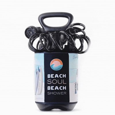 Photograph of a black portable Beach Soul Beach Shower unit with coiled hose and shower head, featuring a light blue label with an orange wave logo, text, and illustrations of surfboards and a beach hut