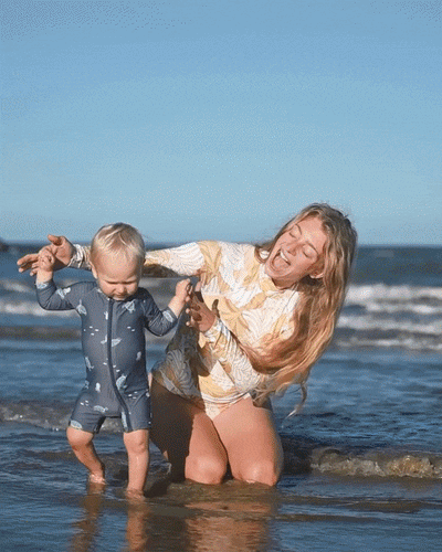 Photograph of a laughing mother in a yellow leafy rash guard and her baby in a grey turtle-patterned rash guard playing in shallow beach water