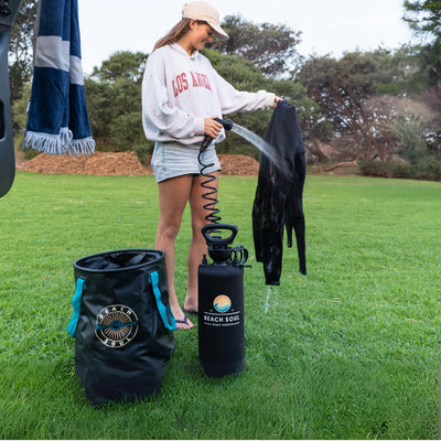 A young woman in a beige cap and Los Angeles sweatshirt rinses a black wetsuit with a Beach Soul portable shower next to a matching bucket with teal handles on a grassy outdoor area