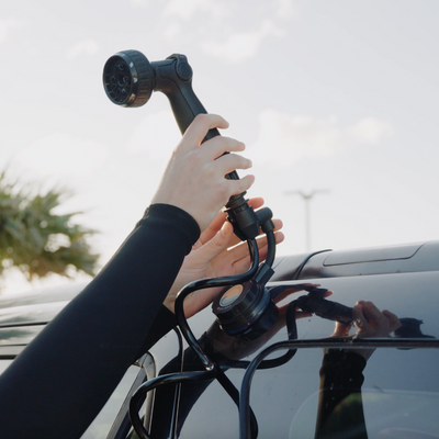 Hands in a black long-sleeved top attaching a black portable shower system featuring a circular Beach Soul logo with a blue wave and orange sun to a dark car roof rack beneath a bright sky with a distant palm tree