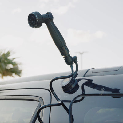 Black portable shower head with multi-pattern nozzle attached by coiled hose and blue and yellow logo suction mount to a dark vehicle's window frame against a bright sky with a blurry palm tree
