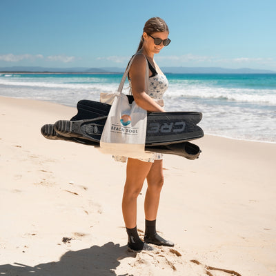 Photography of a smiling woman in a floral dress, carrying a Beach Soul tote bag and black CRESSI fins, on a sunny turquoise beach