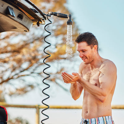 Photograph of a smiling man showering outdoors from a portable car-mounted shower, catching water in his hands, wearing colorful striped swim shorts against a blurred tree background