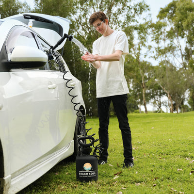 A young man in glasses and a white t-shirt washes his hands with a stream of water from a black Beach Soul portable shower unit featuring a sunset logo and coiled hose, set next to a white car with an open trunk on vibrant green grass in a park-like outdoor area