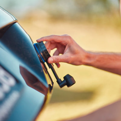 Close-up of a person's hand plugging a black charger with a blue indicator light into a dark vehicle's port against a soft golden outdoor backdrop