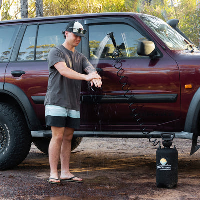 A man rinses his hands with a black Beach Soul portable shower unit beside a dark red 4x4 vehicle featuring a snorkel and rugged mud-terrain tires in a natural wooded outdoor setting