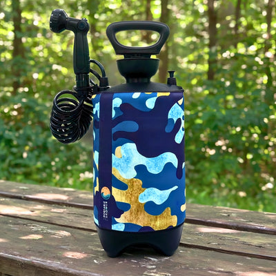 A portable sprayer with a blue light blue and gold abstract camouflage patterned sleeve displaying a "Portable Shower" brand logo and a sun/wave emblem, a black coiled hose, and adjustable nozzle sits on a weathered wooden picnic table outdoors against a blurred green forest backdrop