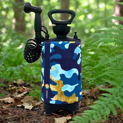 A black portable shower unit with a blue and gold camouflage patterned sleeve, featuring a black coiled hose and shower head, rests on a sun-dappled forest floor among lush green ferns and fallen leaves