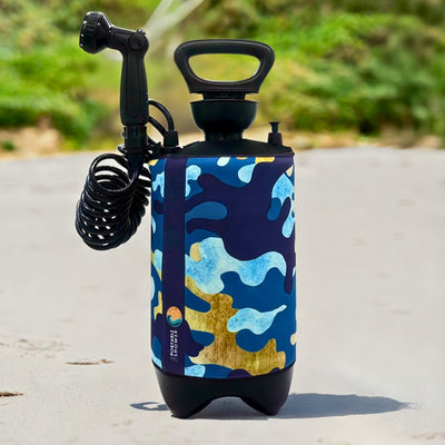 A portable shower with a vibrant blue, light blue, and gold camouflage pattern stands on a sandy beach, featuring a black showerhead spraying water and a coiled hose