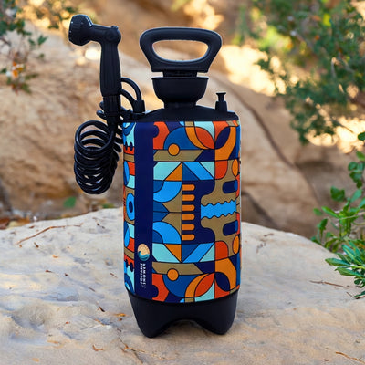 Product photography of a portable shower with a vibrant blue, orange, and brown abstract geometric pattern, featuring a black handle, shower head, coiled hose, and "Portable Shower" logo, resting on a sandy rock outdoors