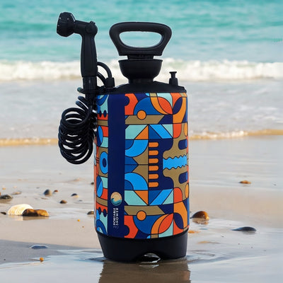 A portable shower with a vibrant geometric pattern in orange, blue, teal, and brown, featuring a black coiled hose and shower head, standing on a wet sandy beach with calm ocean waves and scattered seashells in the background