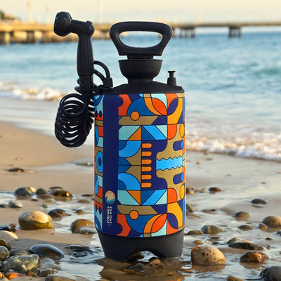 Photography of a portable shower with a vibrant blue orange and teal geometric pattern, black handle, and coiled hose with showerhead, standing on a wet rocky beach with small waves and a distant pier